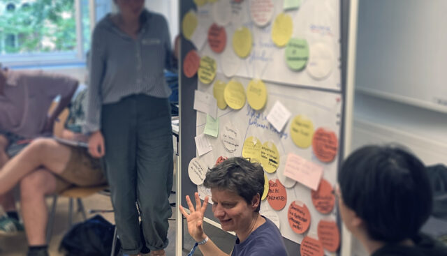 Silja Graupe sitzt auf dem Boden vor einer Pinwand, an welcher Ergebnisse eines 4FutureLabs Workshops festgehalten wurden.
