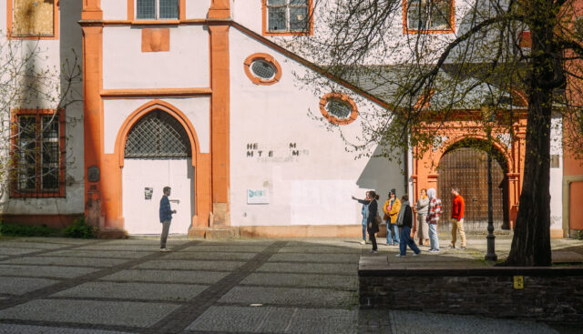 Ein Studierender steht im Rahmen einer Methode zur Zukünftebildung sieben anderen Teilnehmendengegenüber. Die Übung findet vor einem historischen Gebäude in der Stadt Koblenz statt.