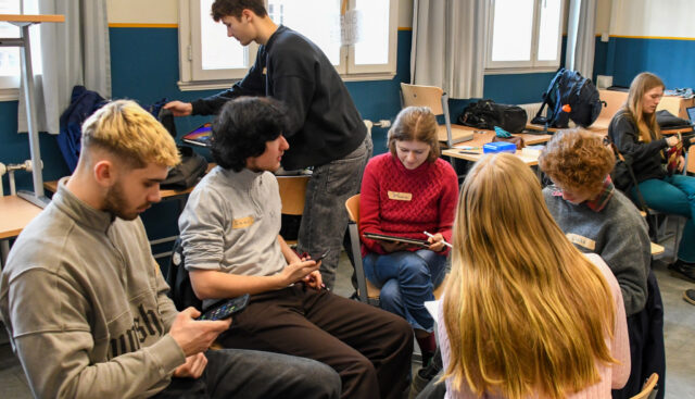 Fünf Schülerinnen und Schüler sitzen in einem Stuhlkreis und imaginieren Zukunftsszenarien. Alle halten hierbei elektronische Geräte in Form von Smartphones und Tablets in ihren Händen.