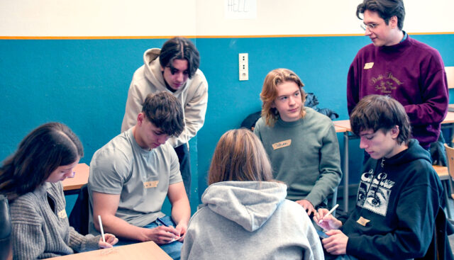 Paul Kühn, wissenschaftlicher Mitarbeiter für Zukünftebildung an der Hochschule für Gesellschaftsgestaltung (HfGG) aus Koblenz steht vor sechs Schülerinnen und Schülern, welchen er eine 4FutureLab Übung anleitet. Die Schülerinnen und Schüler sitzen und machen sich Notizen.