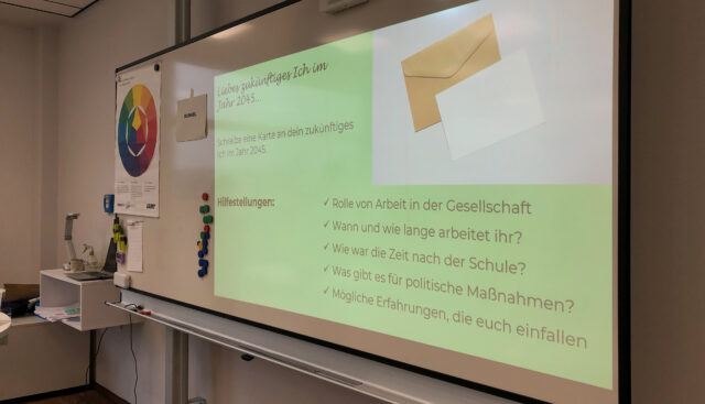 Auf einem Monitor wird eine Übung für Schülerinnen und Schüler erklärt: Sie sollen eine Postkarte an das zukünftige Ich im Jahr 2045 schreiben.